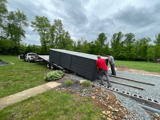 Two men unload large panels from a trailer onto a gravel driveway. Trees in the background.