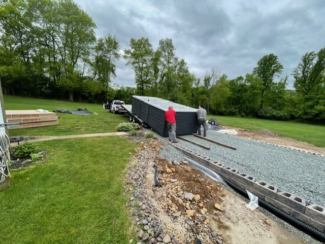 Two men guiding a large, dark rectangular structure being unloaded onto a gravel path outdoors.