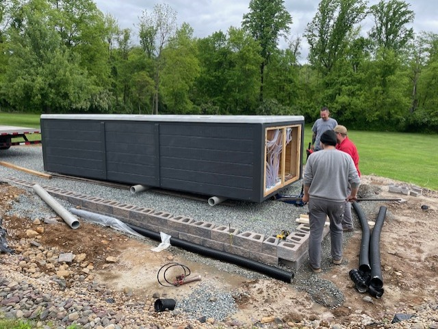 Men installing a dark gray modular building on a gravel pad outdoors, surrounded by construction materials.