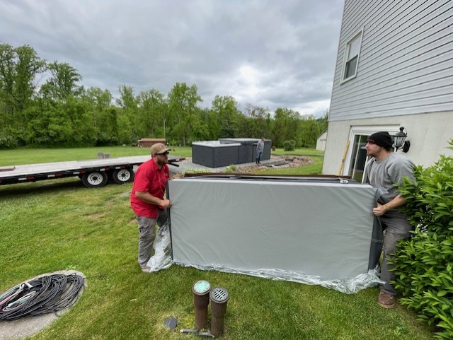 Two men lifting a large, wrapped white panel on a grassy yard near a house. A trailer is parked nearby.