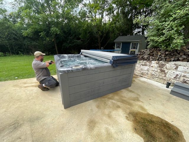 Man installing a grey hot tub on a concrete patio outdoors, near greenery and a small shed.