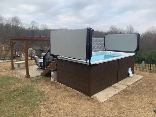 Hot tub with open gray cover, next to a pergola on a grassy hill.
