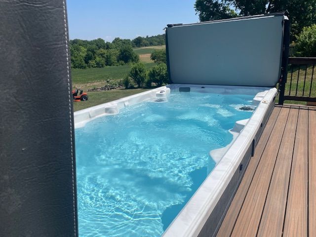 Swim spa on a wooden deck, blue water reflecting the sky, cover open, landscape background.