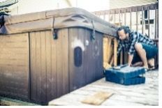 Person crouching beside a hot tub, placing a blue cover on a wooden deck in a backyard.