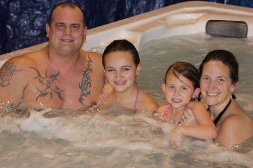 Family of four smiling in a hot tub. Man with tattoos, two girls, and woman are enjoying the warm water.