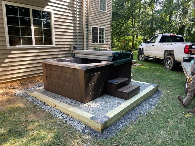 Hot tub on a gravel base next to a house with steps; white truck parked in the background.
