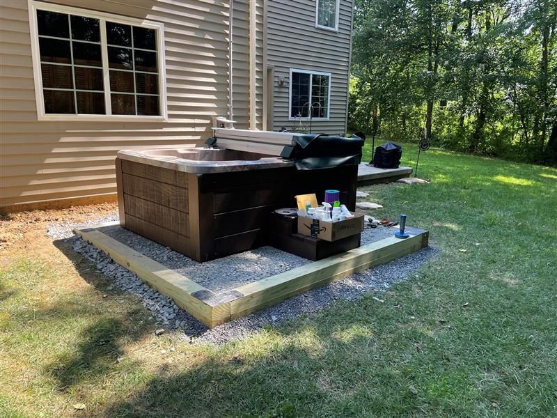 Hot tub on gravel base next to a beige house. Supplies and trash bag sit nearby.