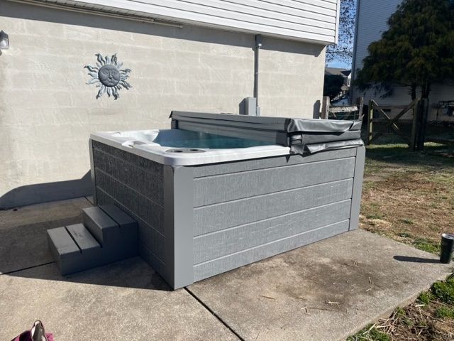 Gray hot tub on a concrete patio with a set of steps, next to a light-colored wall.