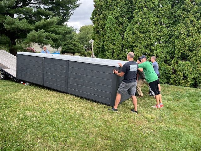 Four people lifting a dark gray hot tub from a trailer onto a grassy yard.