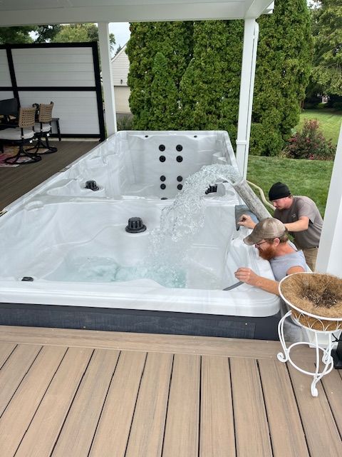 Two men working on a large white hot tub on a wooden deck, under a covered area. Water is visible.