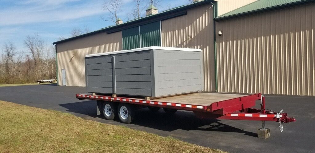 Flatbed trailer carrying two gray portable toilets, parked in front of a large tan industrial building.