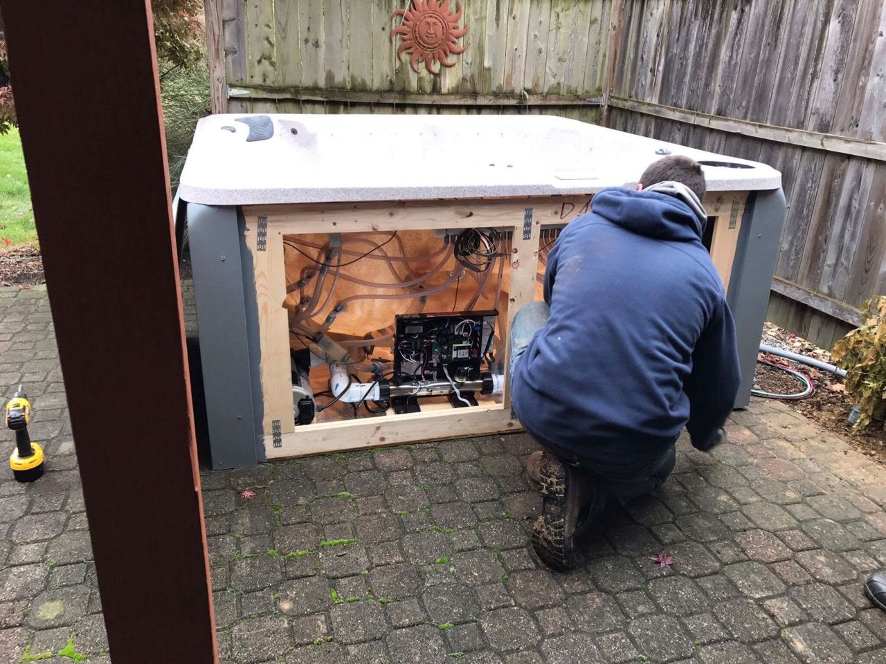 Man kneels working on hot tub machinery outdoors. The hot tub is gray with its side open to the components.