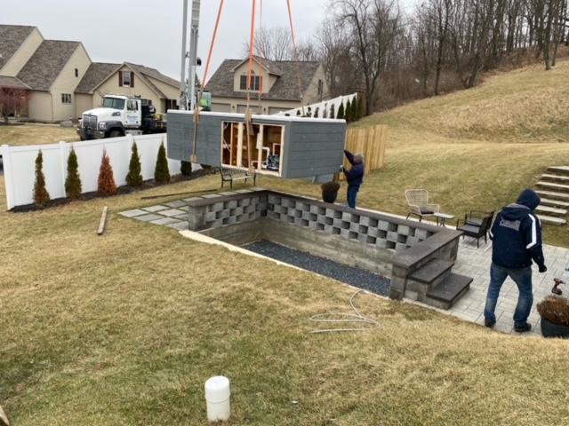 A container is being lowered into an empty pool by a crane; workers observe on a grassy hill near homes.
