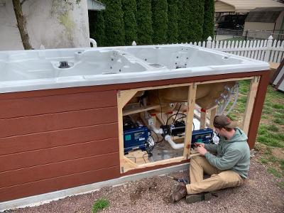 Man working on the mechanics of a hot tub's frame, with a red-brown panel, outside.