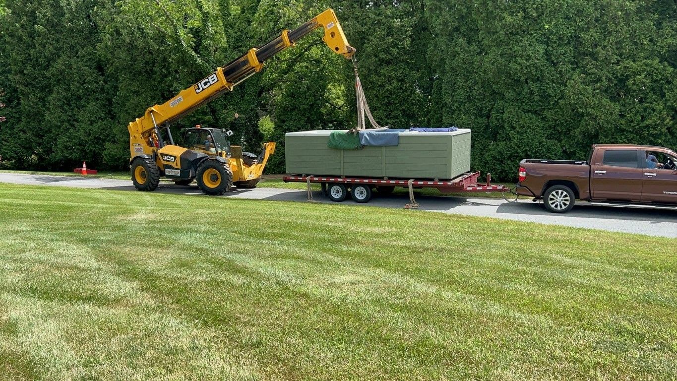 Yellow lift crane lifting a green hot tub off a trailer pulled by a brown pickup truck on grass.