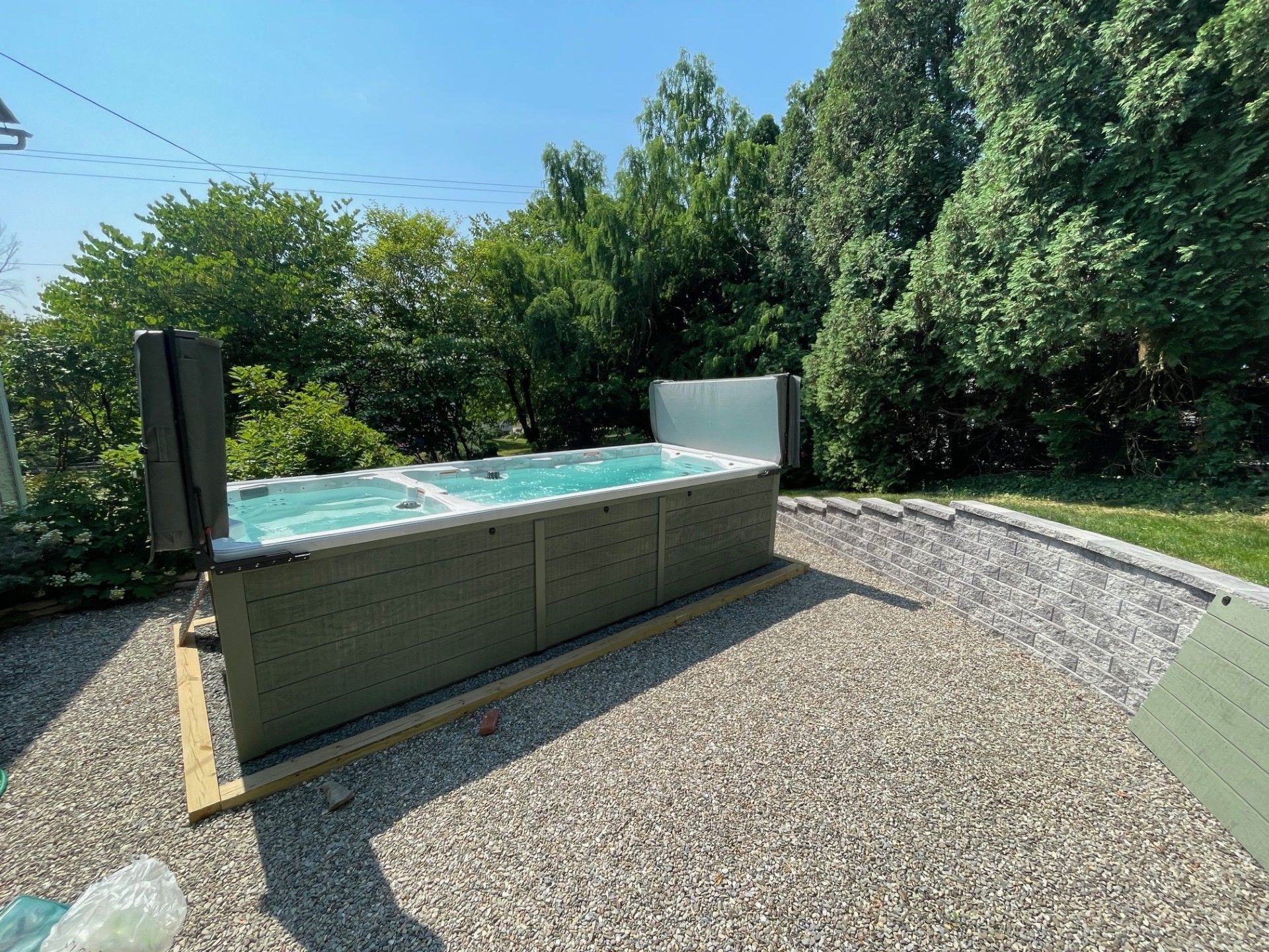 A rectangular spa with open covers sits on gravel outdoors, surrounded by trees and a retaining wall.