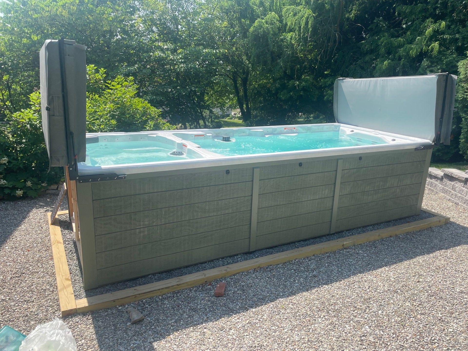 Outdoor hot tub with open lid on gravel, surrounded by trees.