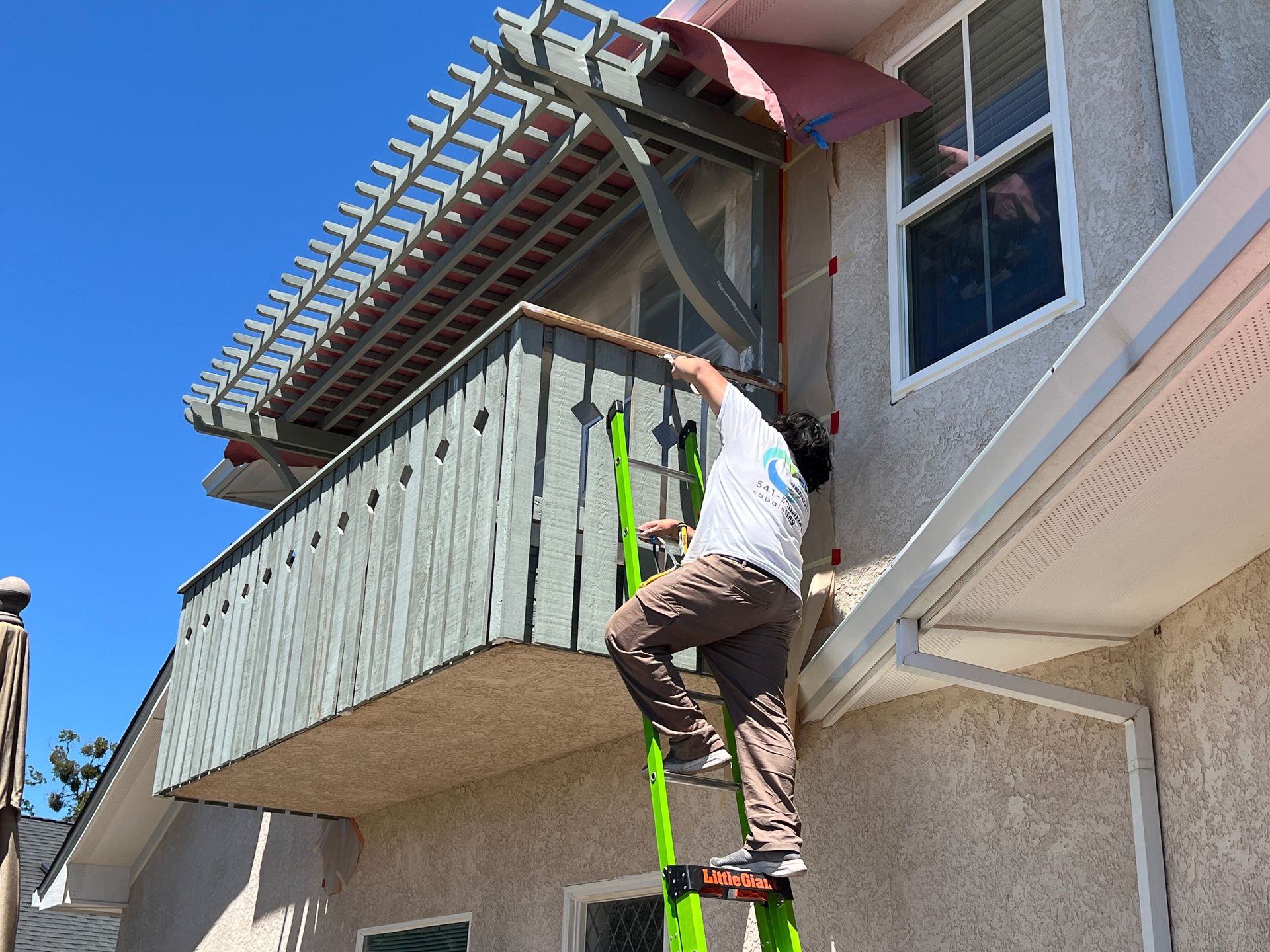 Man on ladder, working on a house exterior.  He is reaching towards a window with a balcony.  Sunny, outdoor setting.
