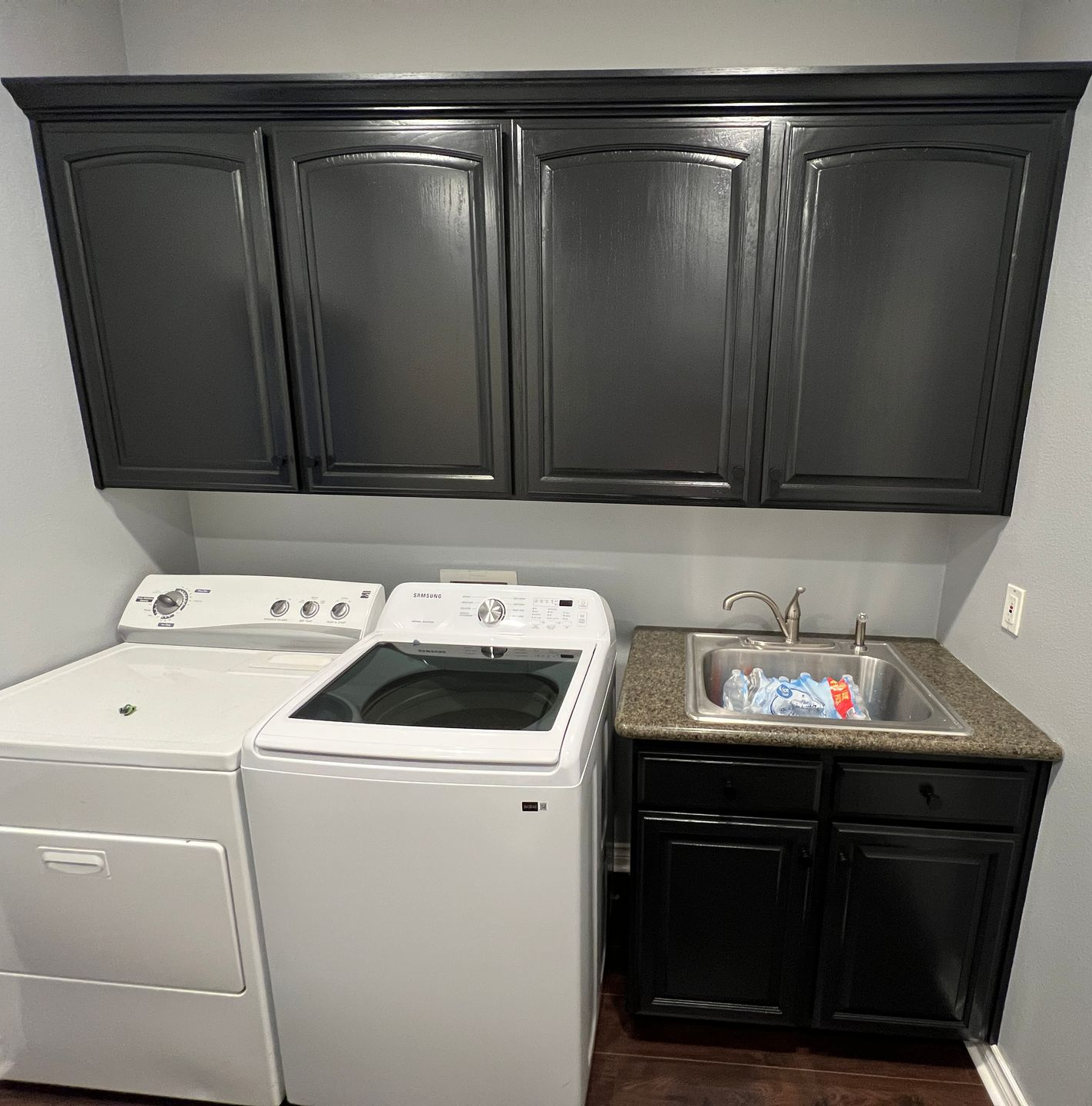 Laundry room with black cabinets, white appliances, and a sink. Gray walls and a white door are visible.