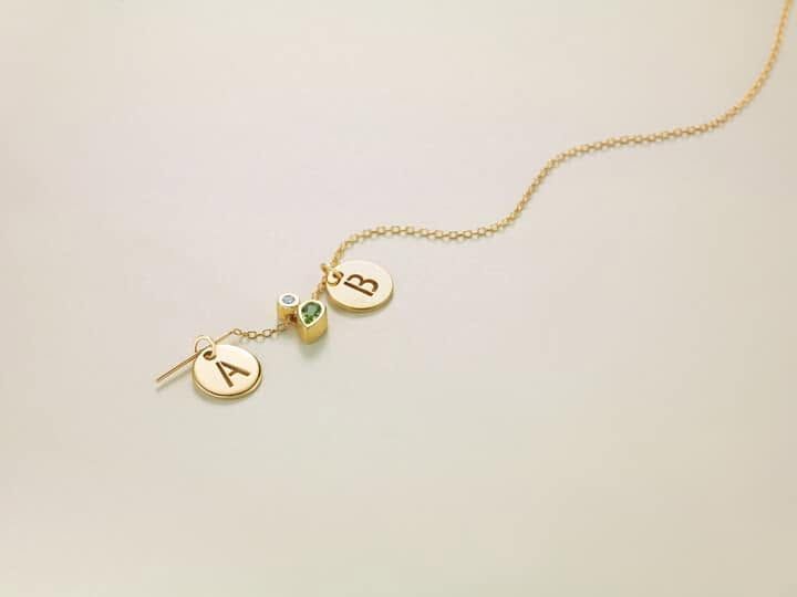 Gold necklace with personalized letter charms and a small green gemstone on a beige background
