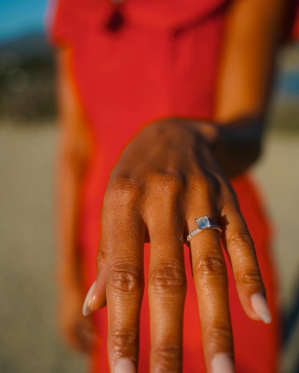 Close-up of a hand with a diamond ring, wearing a red dress against a blurred outdoor background
