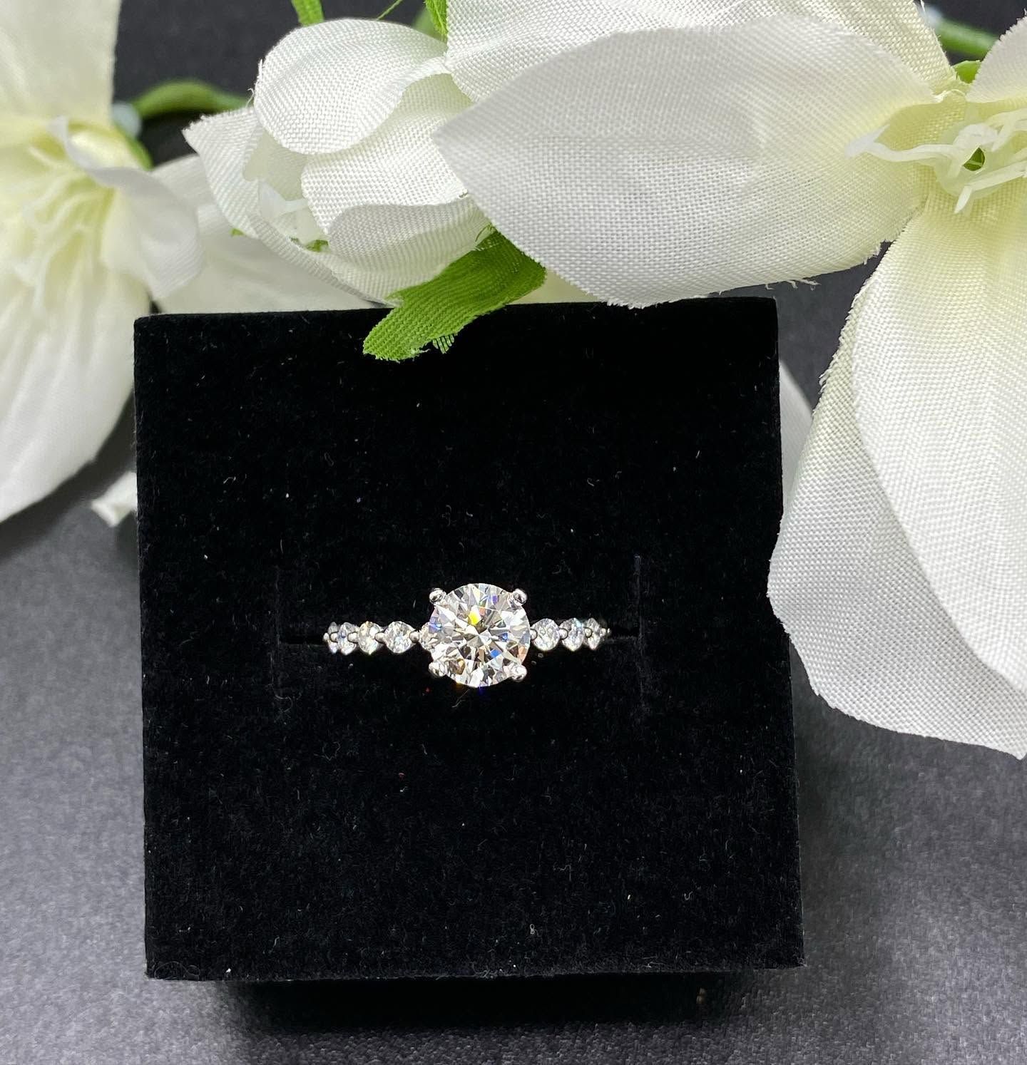 Diamond ring on a black display stand, framed by white flowers.