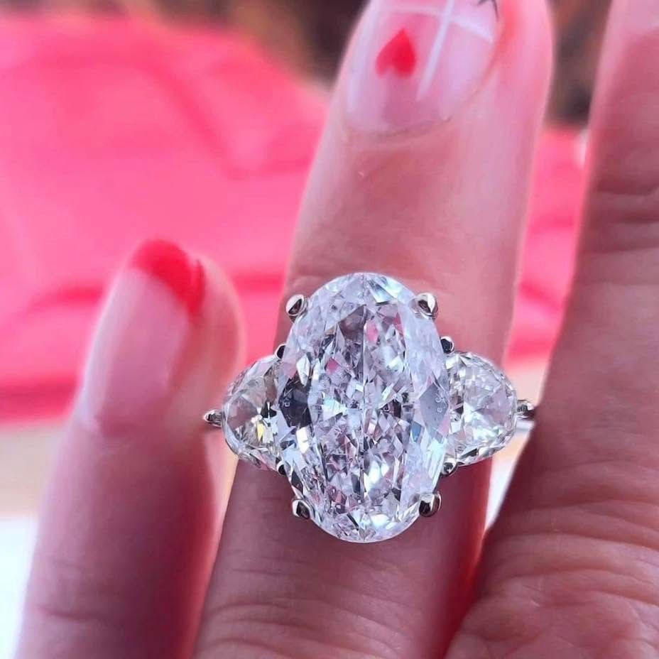 Close-up of a hand wearing a large oval diamond ring with side stones on a pink background