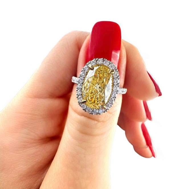 Hand with long red nails holding a large yellow diamond ring with a halo setting