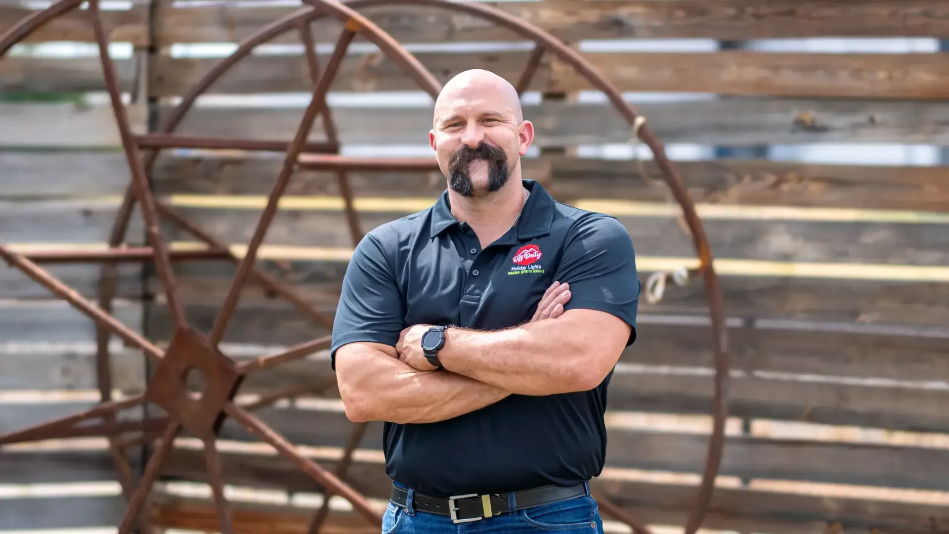 Man with crossed arms standing in front of a rustic wooden wall and metal wheel structure