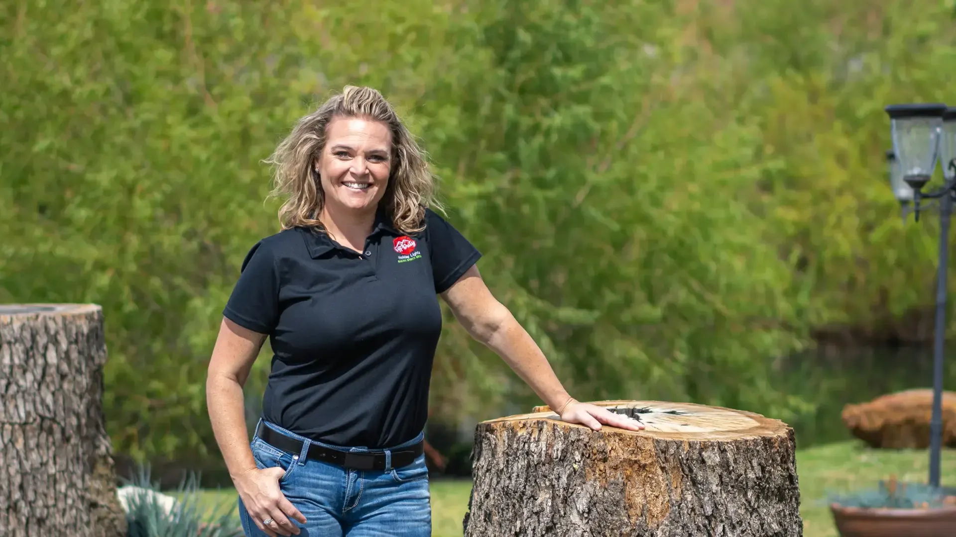 Woman in black polo and jeans standing by tree stump in a green outdoor setting