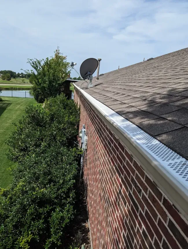 Brick house roofline with satellite dish, white gutter, and green shrubs beside a pond