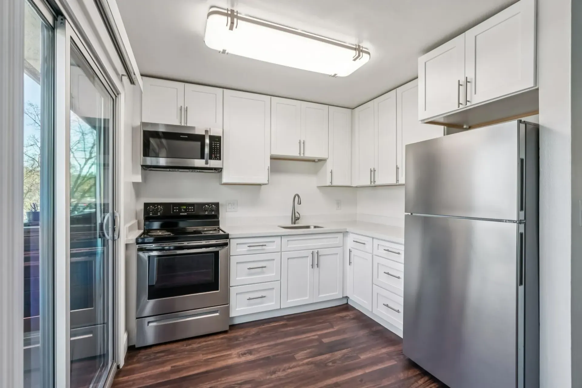 Bright white kitchen with stainless steel fridge, oven, microwave, and a sliding glass door on the left.