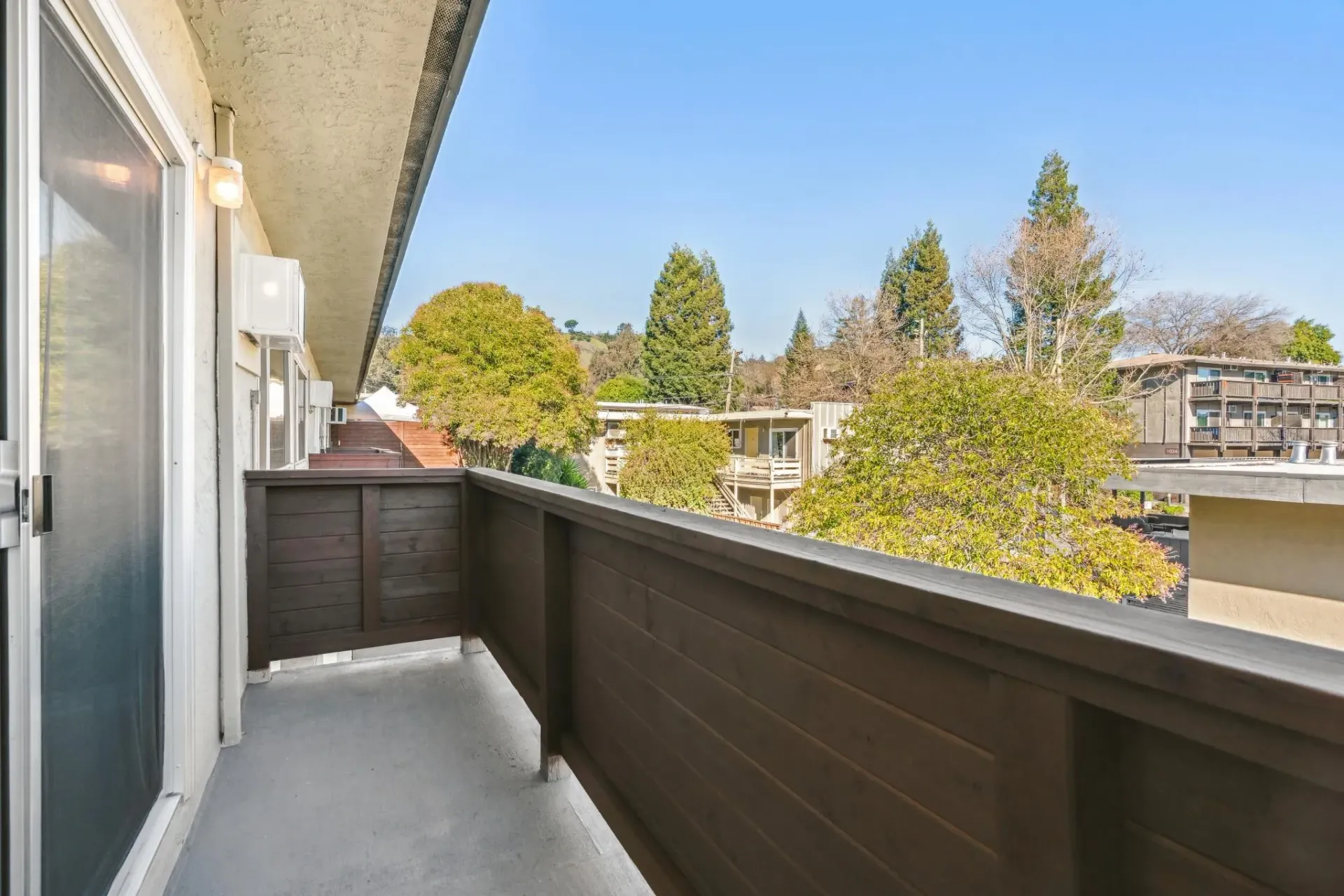 Balcony view from an apartment with a brown railing, sliding door, and trees and neighboring buildings in the distance.
