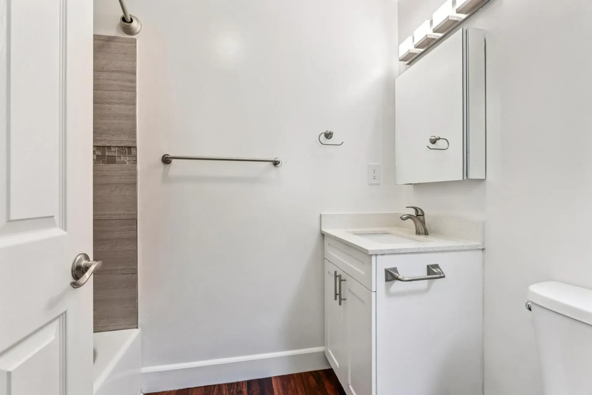 Apartment bathroom with white vanity, mirror cabinet, towel bar, toilet, and shower.
