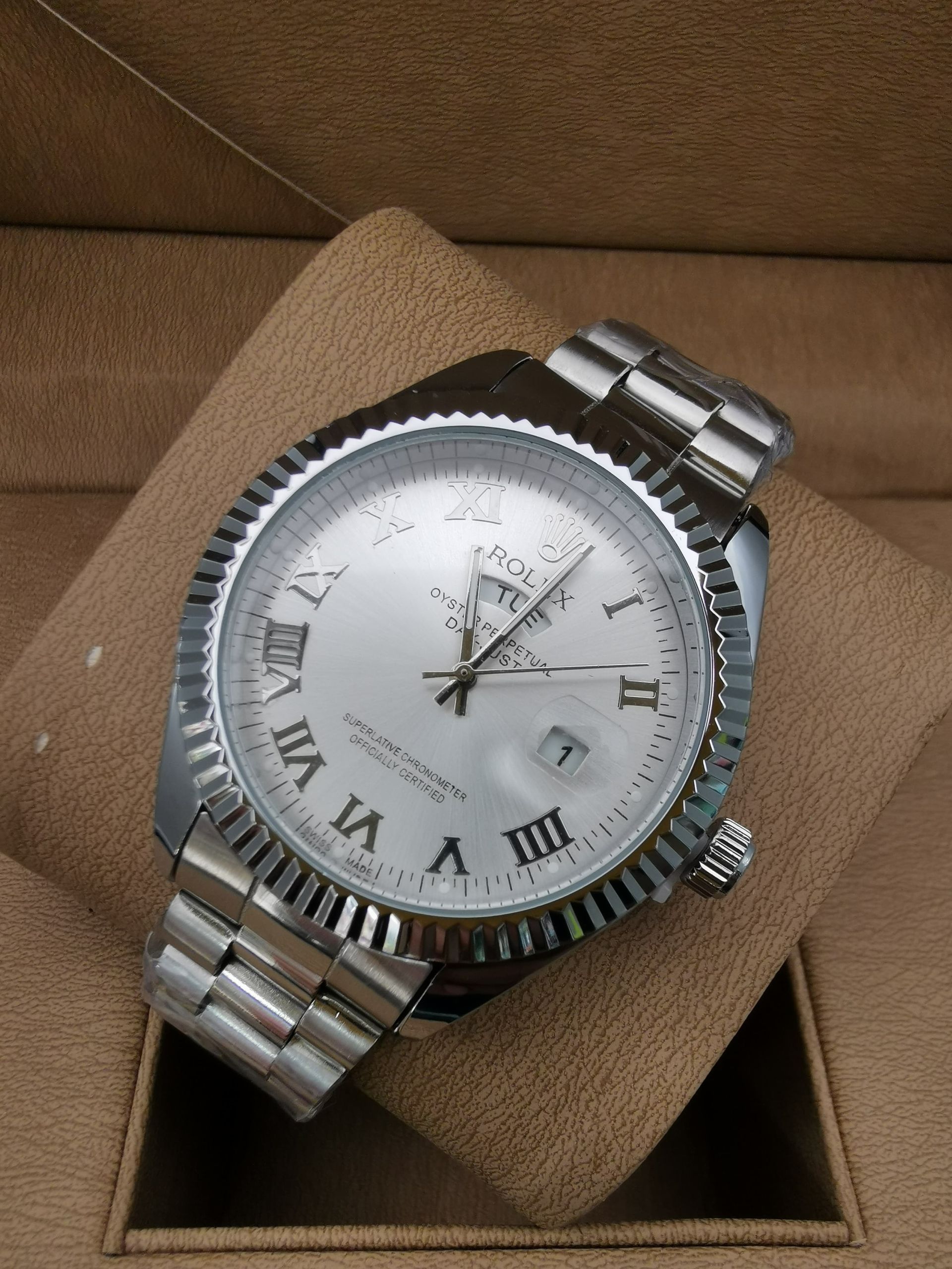 Reloj de pulsera plateado con esfera blanca y números romanos, sobre una almohada marrón en una caja.