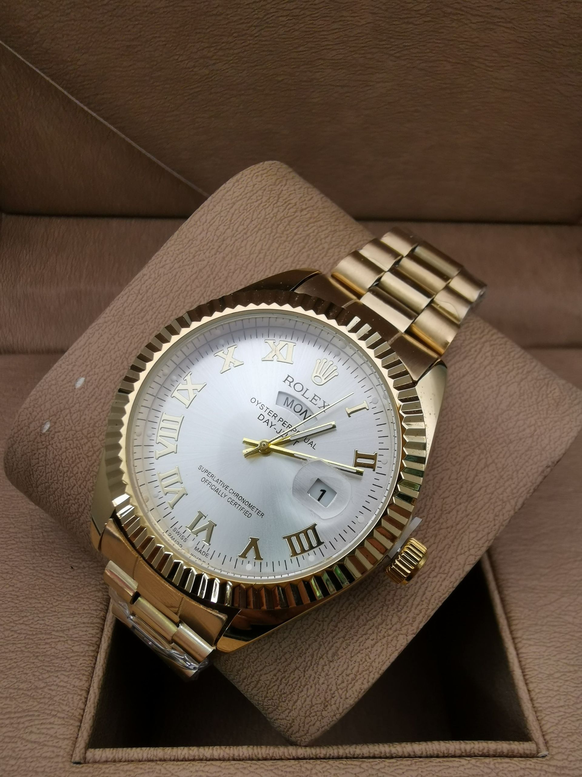 Reloj de pulsera plateado con esfera blanca y números romanos, sobre una almohada marrón en una caja.