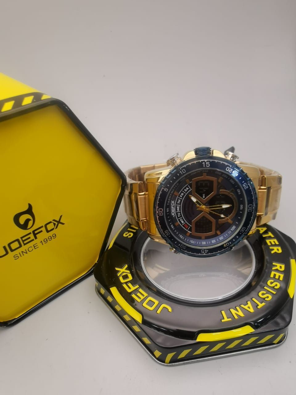 Reloj JOEFOX dorado sobre expositor negro y dorado. Caja amarilla con el logotipo al fondo.
