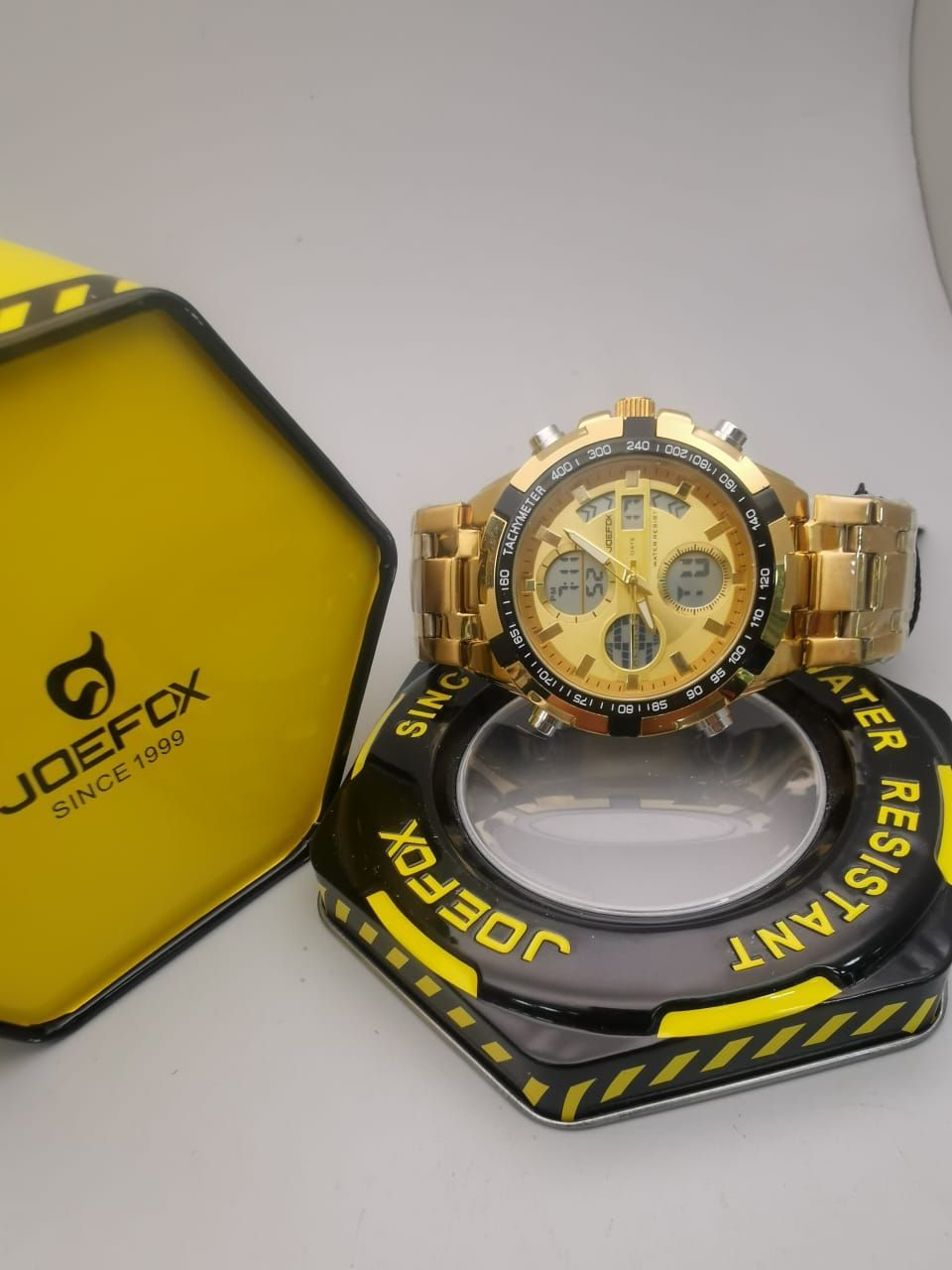 Reloj JOEFOX dorado sobre expositor negro y dorado. Caja amarilla con el logotipo al fondo.