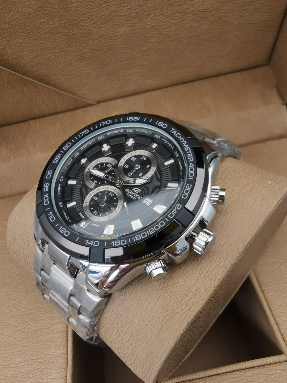 Reloj de pulsera plateado con esfera negra y funciones de cronógrafo, apoyado sobre un cojín marrón.