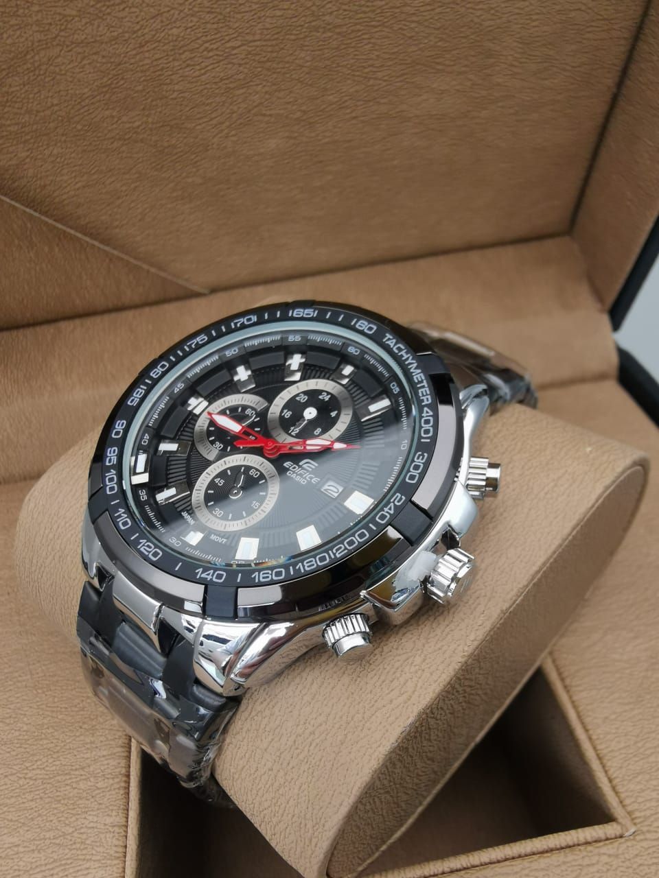 Reloj de pulsera plateado con esfera negra y funciones de cronógrafo, apoyado sobre un cojín marrón.