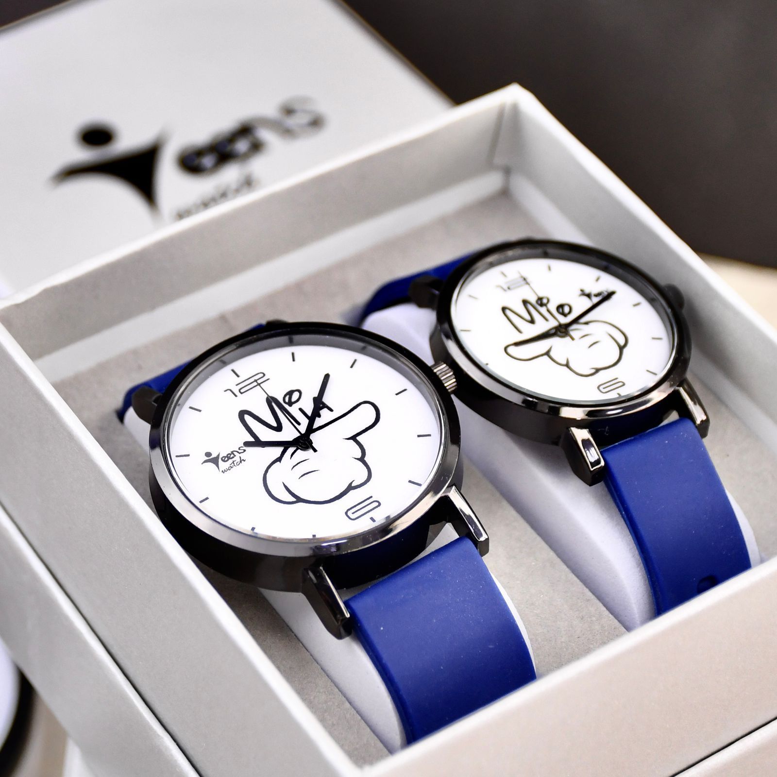 Par de relojes en una caja de regalo, cada uno con cara blanca, correa azul y diseño de manecillas de dibujos animados.
