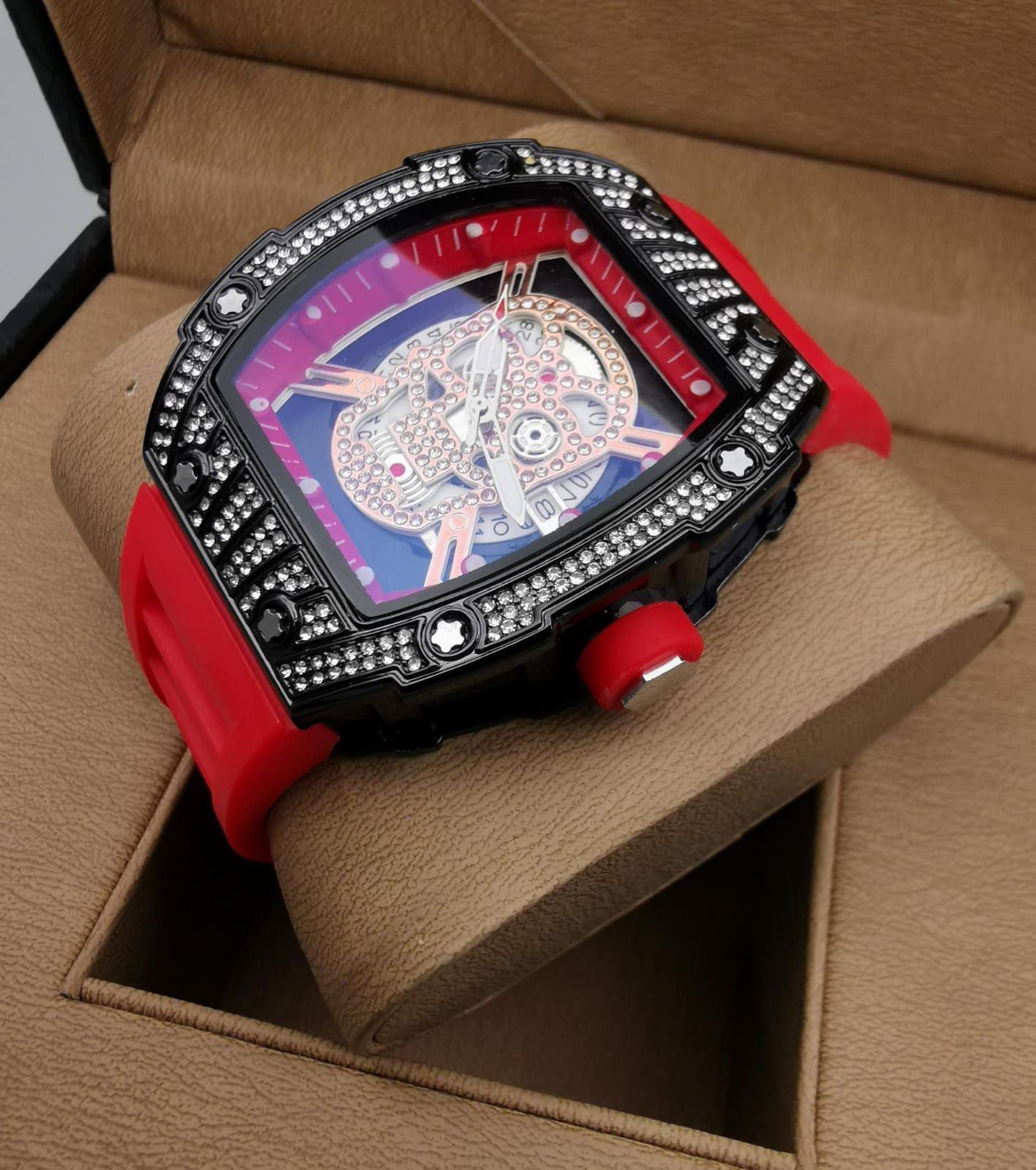 Reloj de pulsera rojo y negro con detalles de joyas, exhibido en un soporte de reloj marrón dentro de una caja.