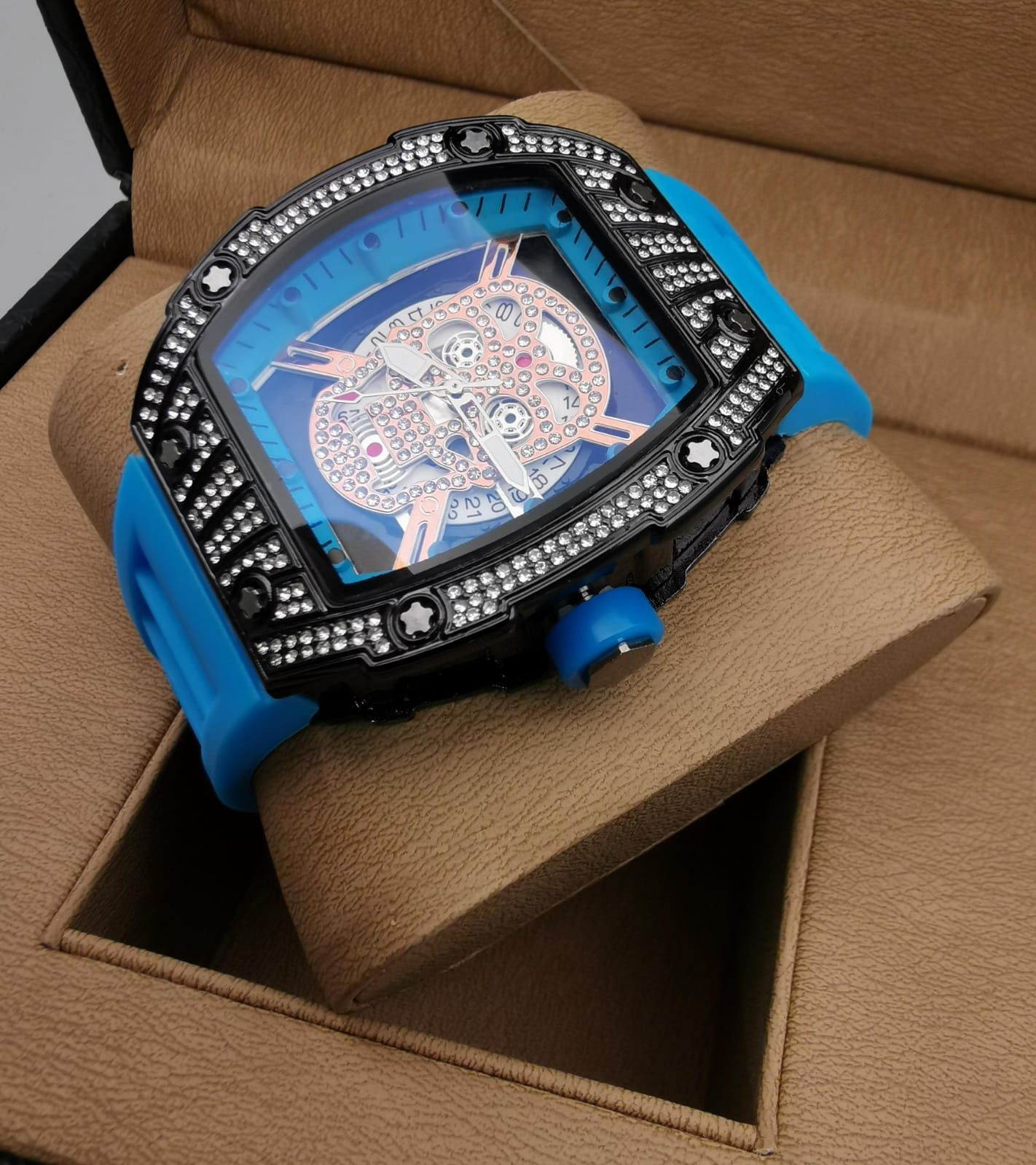 Reloj de pulsera rojo y negro con detalles de joyas, exhibido en un soporte de reloj marrón dentro de una caja.
