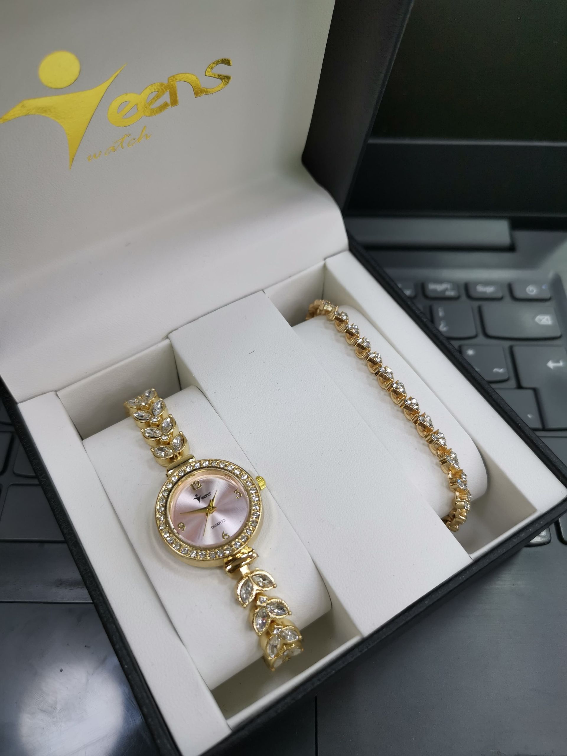 Conjunto de reloj y pulsera de oro en caja negra, con esfera roja y detalles brillantes.