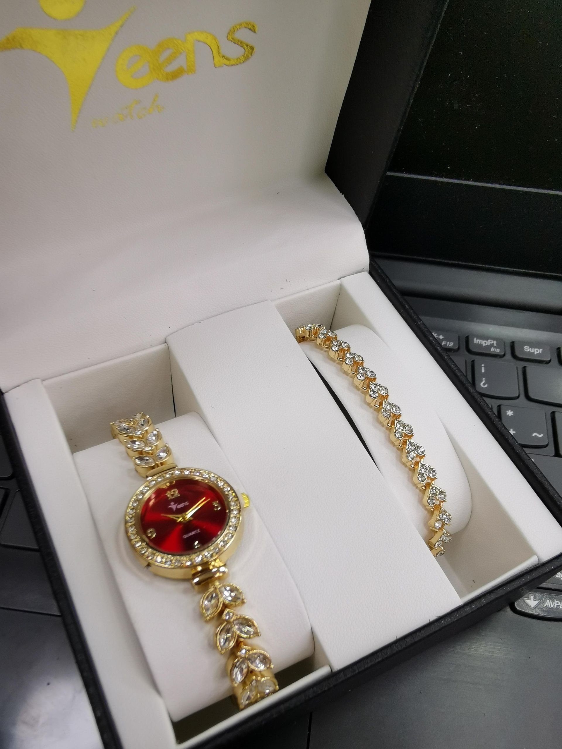 Conjunto de reloj y pulsera de oro en caja negra, con esfera roja y detalles brillantes.