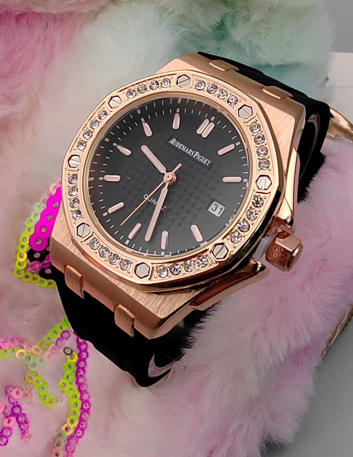 Reloj de oro rosa con esfera negra, bisel de diamantes y correa negra, que reposa sobre un material suave y pastel.