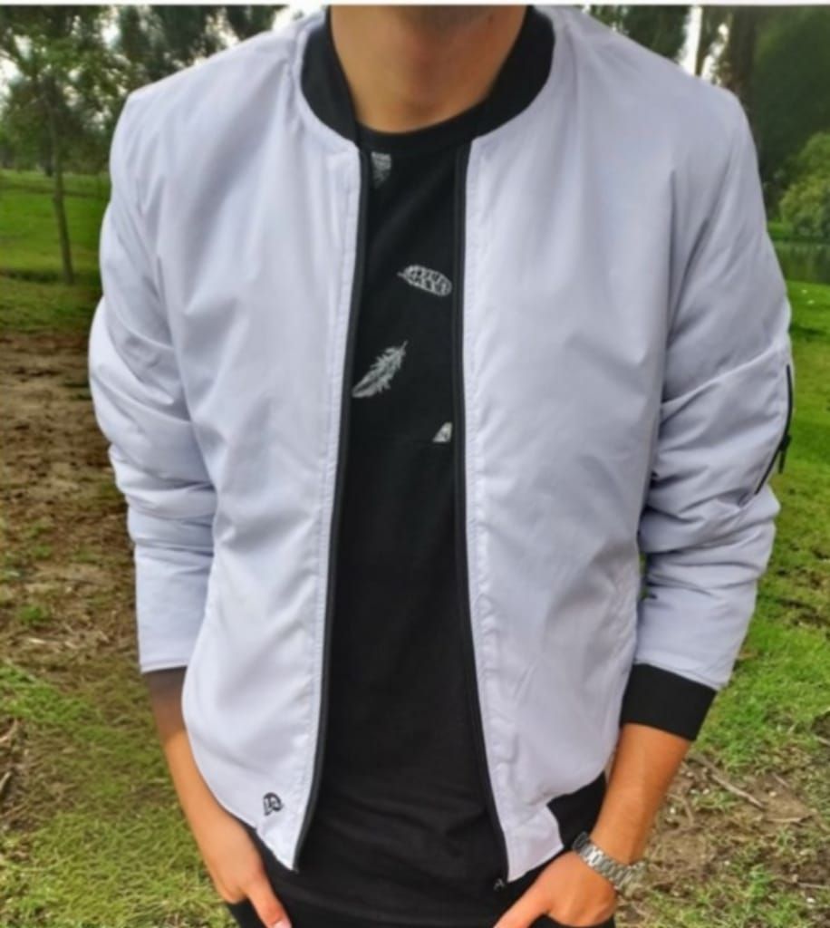 Hombre con chaqueta bomber gris claro sobre camisa negra, manos en los bolsillos, al aire libre.