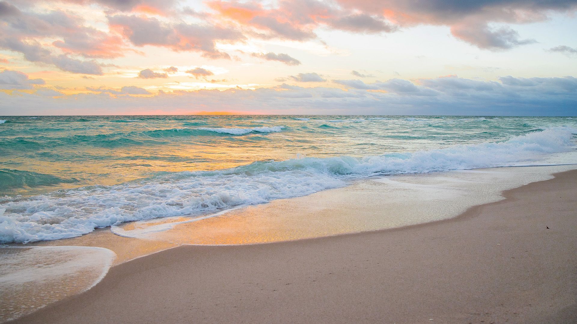 Sandy beach at sunset with turquoise waves and pastel clouds over the ocean