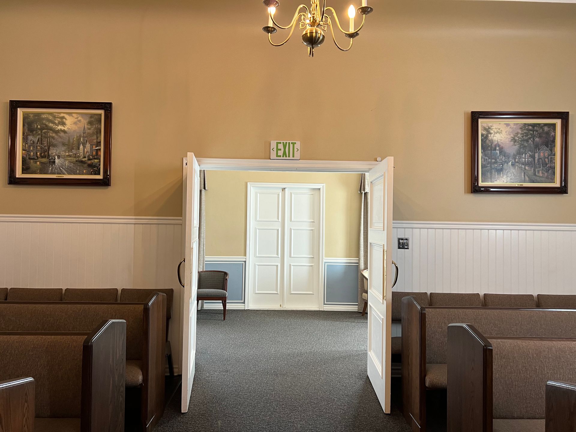 Empty church hall with wooden pews, chandelier, and open double doors leading to another room