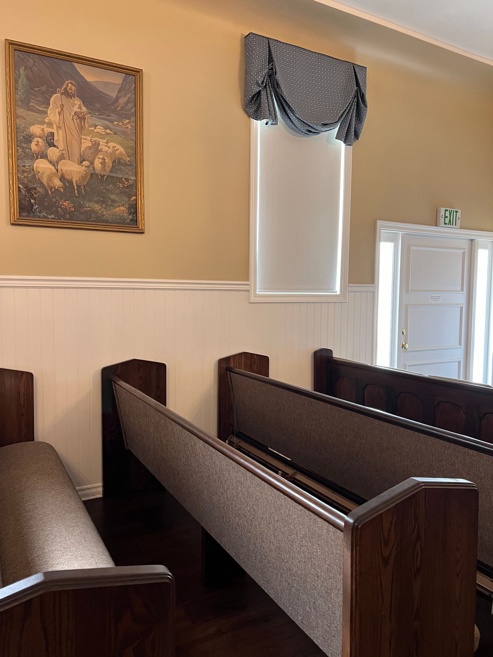 Church pews in a small chapel with beige walls, framed art, and a window with a blue valance.